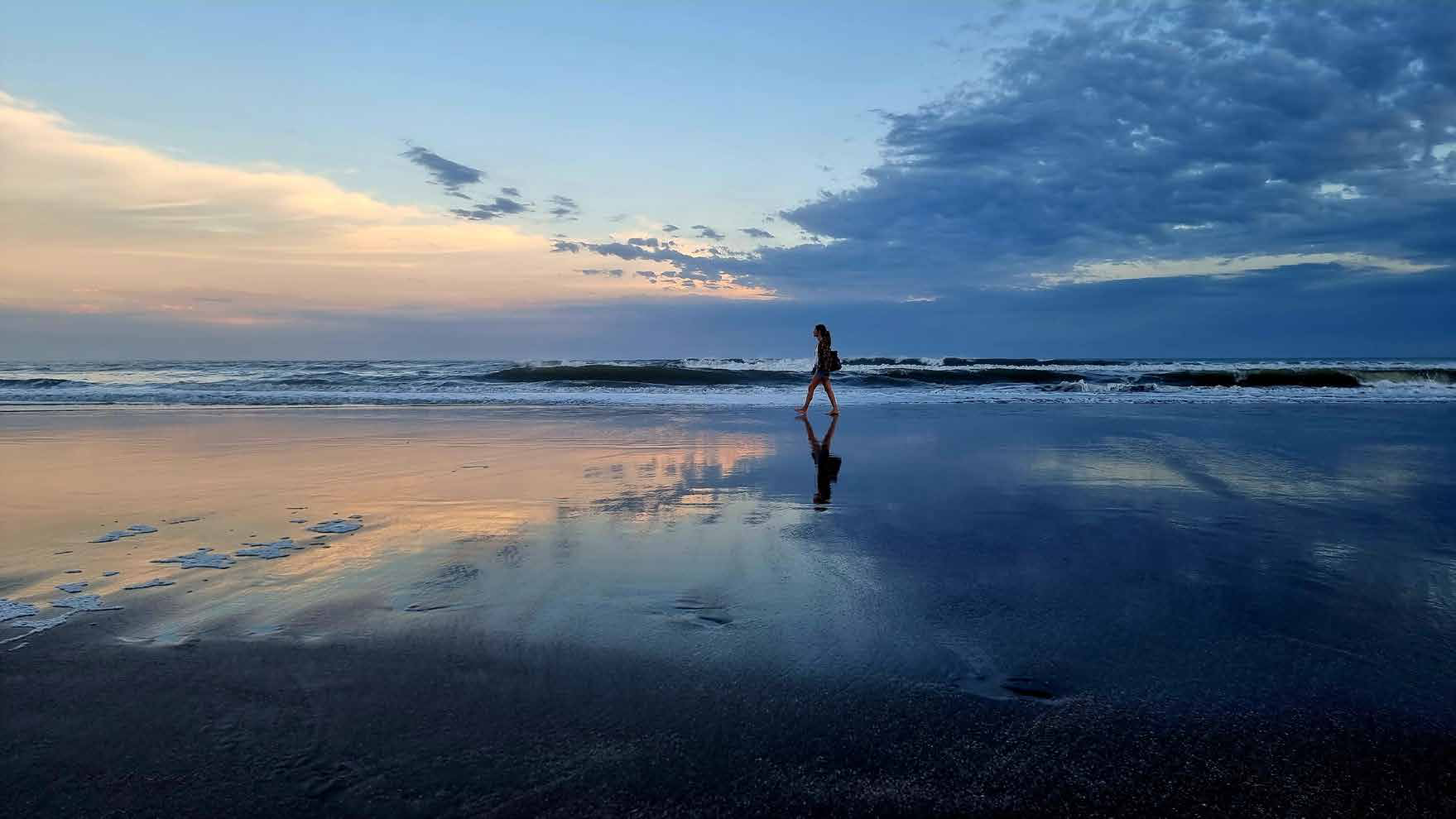 A figure walking the reflective wet sand at blue hour on a dusk beach